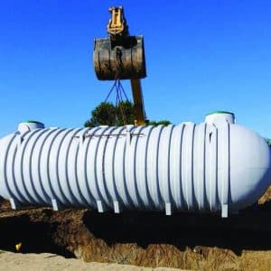 The image shows a large underground cylindrical cistern tank lifted by a crane for installation.